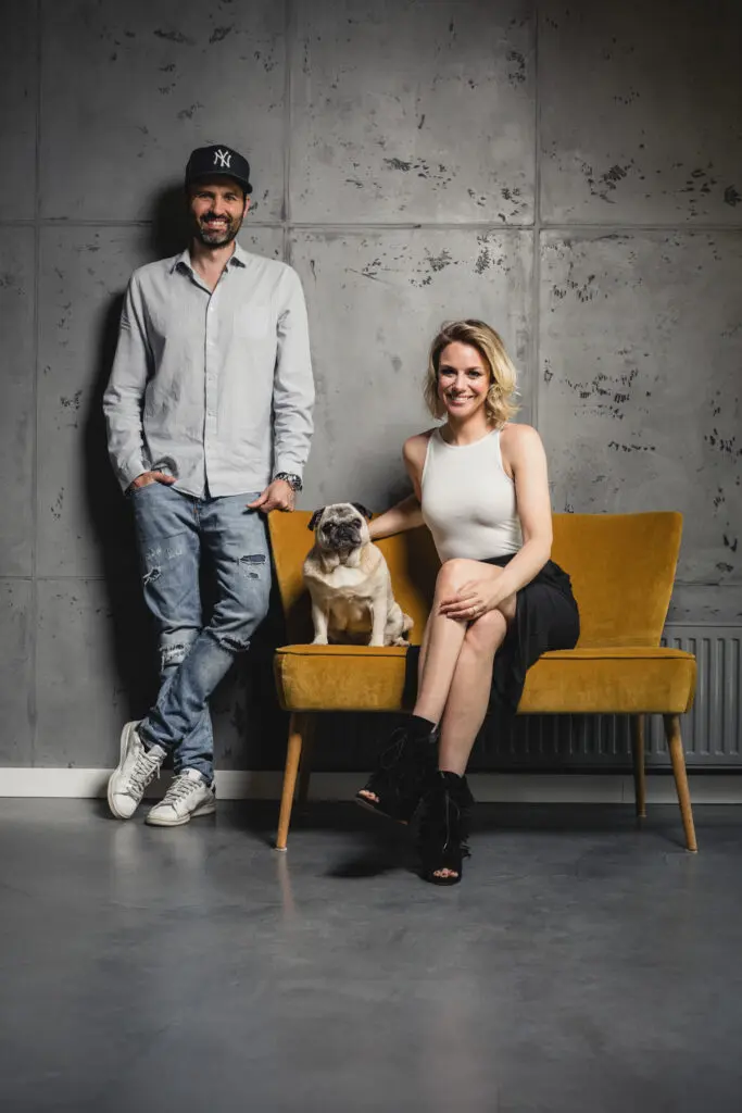 Ein Paar sitzt mit einem Hund auf einem gelben Sofa vor einer Betonoptik-Wand im Wohnzimmer, die stilvolle Wandgestaltung zeigt.
