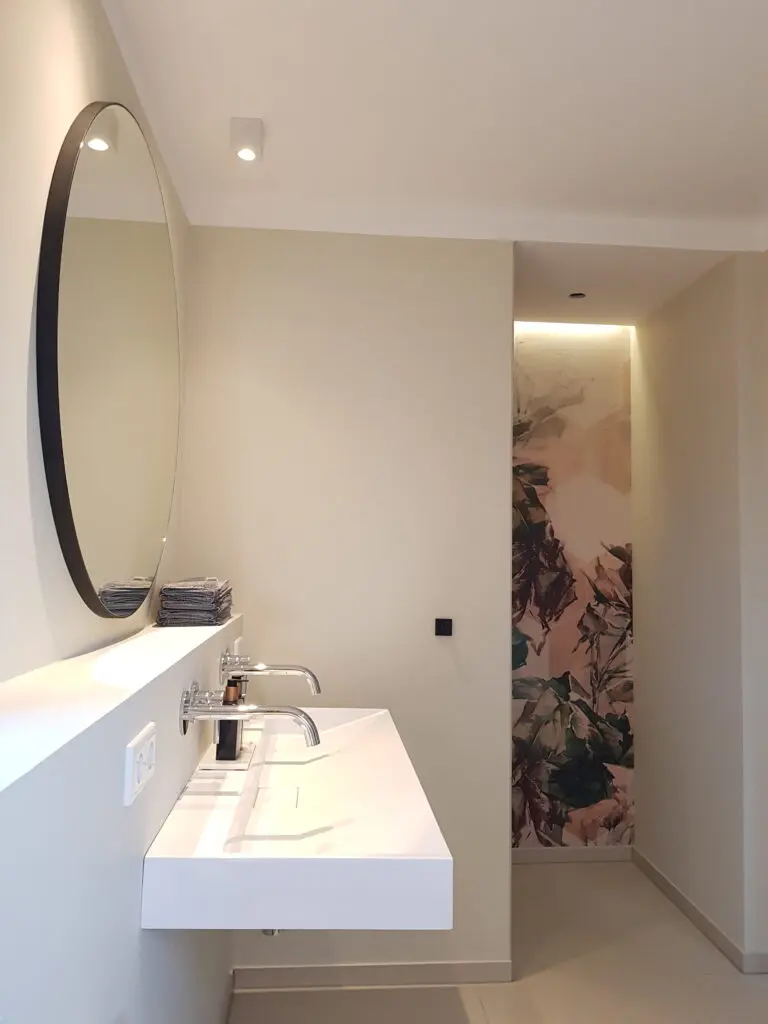 Bad mit Rinnenwaschbecken, Rundspiegel und hinterleuchteter Wandfläche mit rosa Blätterdruck
