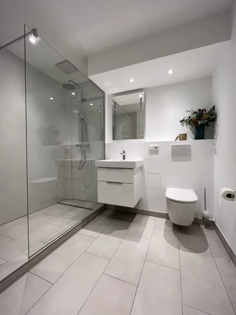 Helles Bad mit fugenloser Walk-in-Dusche, weißem Waschtisch und Hänge-WC auf grauen Bodenfliesen