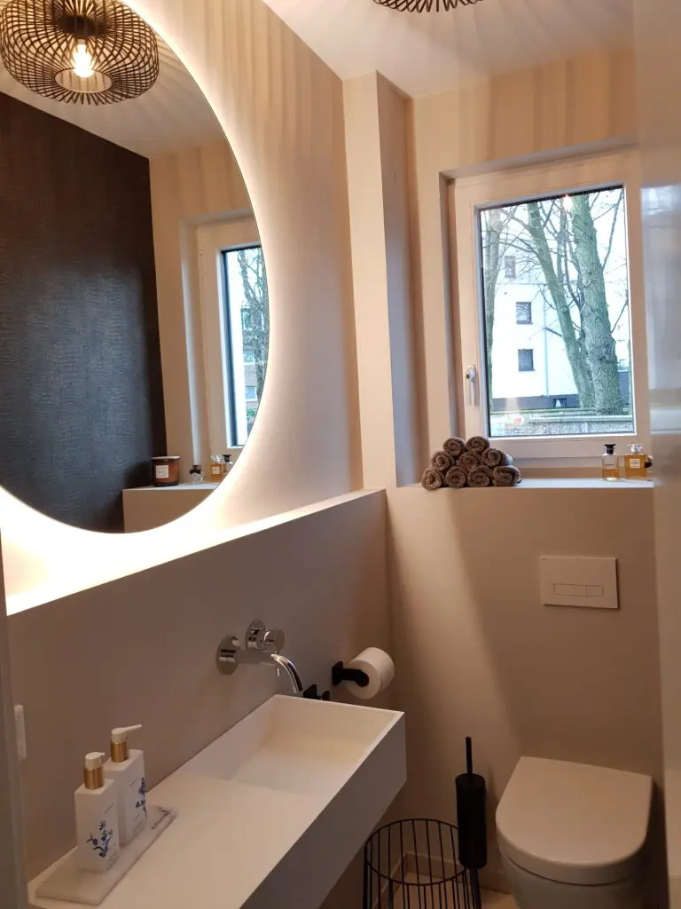 Gäste-WC mit beigen fugenlosen Wänden, Rinnenwaschbecken, rundem beleuchtetem Spiegel und Fenster