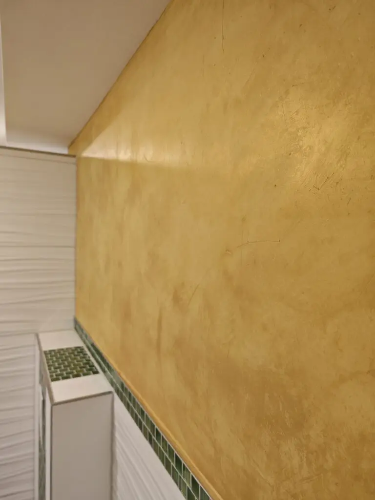 Goldgelb schimmernde Spachtelwand über Wellenfliesen und grünem Mosaikstreifen in Nahaufnahme