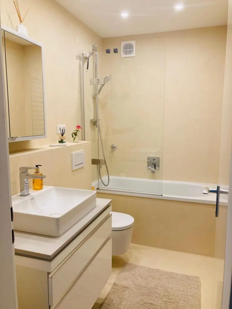 Bad in hellem Sandton mit fugenlosen Wänden, Badewanne mit Glasfaltwand und Waschtisch