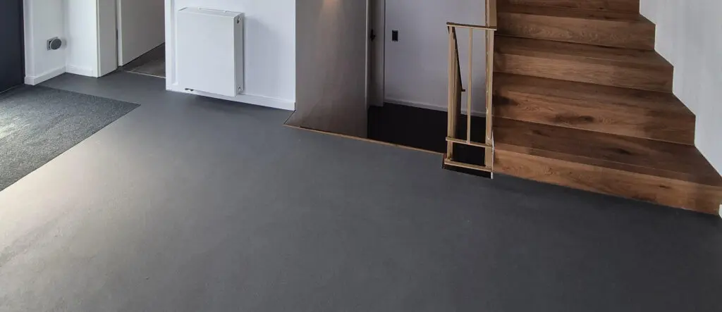 Ein modernes Wohnzimmer zeigt eine graue Bodenbeschichtung in Betonoptik neben einer hölzernen Treppe.