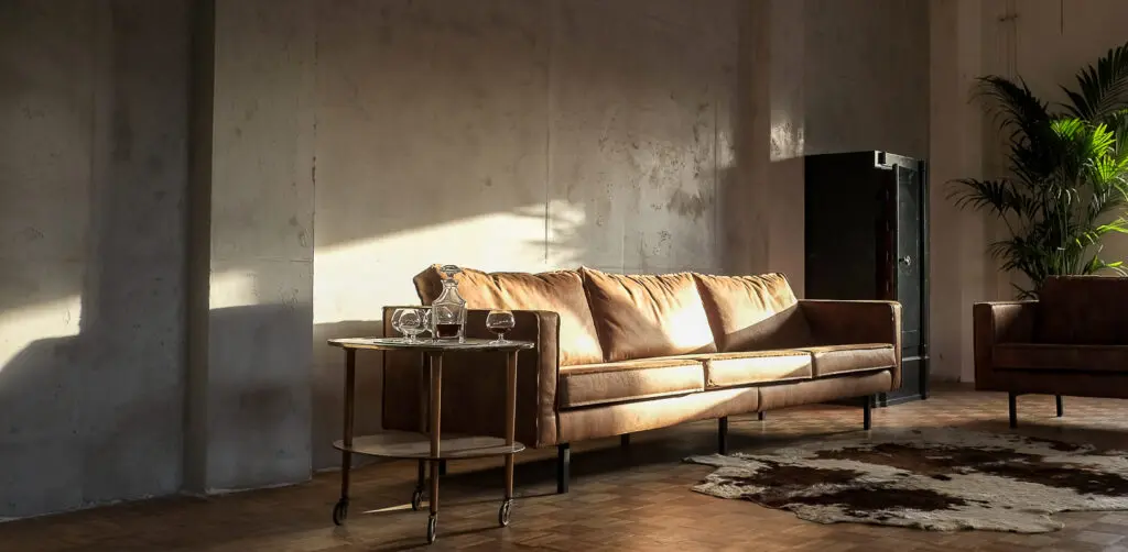 Ein stilvolles Wohnzimmer mit einer Wand in Betonoptik und einem gemütlichen Ledersofa im warmen Sonnenlicht.