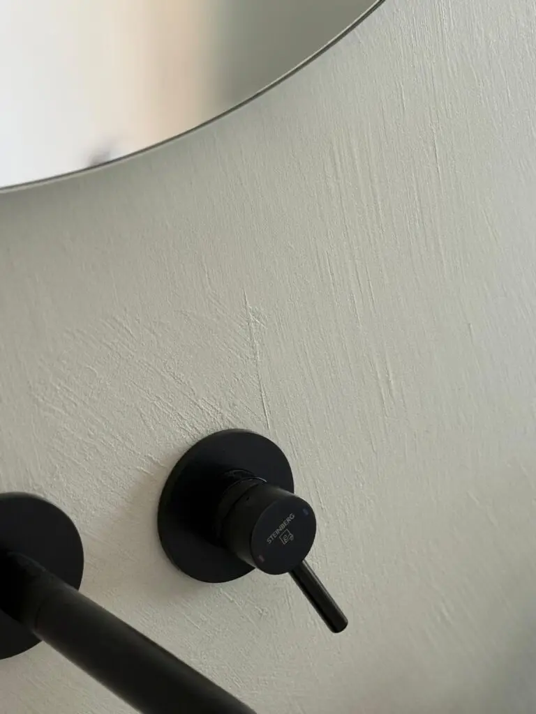 Nahaufnahme hell gespachtelter Wand mit Kellenstruktur, schwarzer Wandarmatur und Spiegelrand