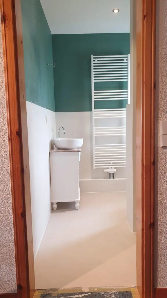 Blick durch Holzzarge in schmales Bad mit grüner Wand, weißem Waschtisch und hellem Boden