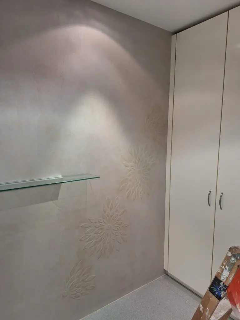 Hellgraue Spachtelwand mit erhabenen Blütenornamenten neben weißem Einbauschrank und Glasablage