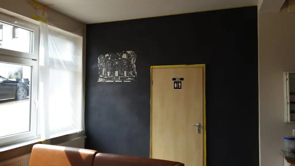 Raum mit anthrazitfarbener Spachtelwand, heller Fassadengrafik und Holztür zum WC