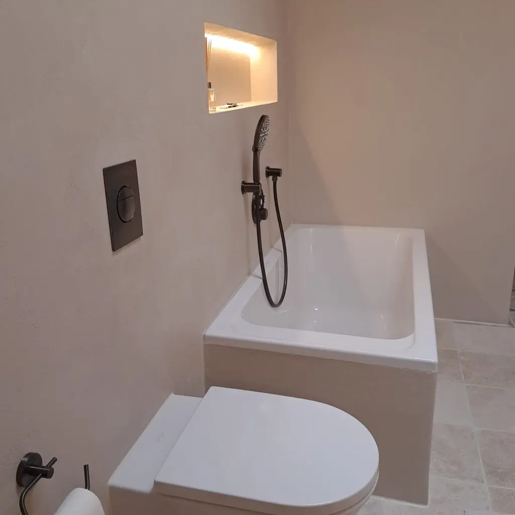 Bad mit hellen fugenlosen Wänden, beleuchteter Nische über Badewanne und schwarzer Handbrause