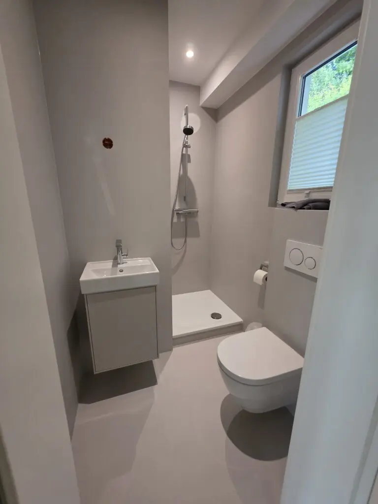 Kleines Duschbad mit hellgrauem fugenlosem Boden, Waschtisch, Duschwanne und Fenster