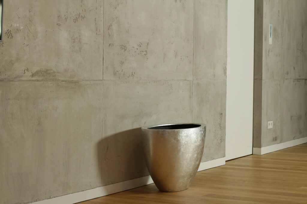 Ein modernes Wohnzimmer zeigt Wände in Betonoptik mit Oberflaeche-STUCCORINO Torino-Beton und einem silbernen Pflanzgefäß.