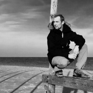 Ein Mann sitzt lässig auf einem Holzzaun am Strand, inspiriert von der kreativen Wandgestaltung von Einhornwerke.