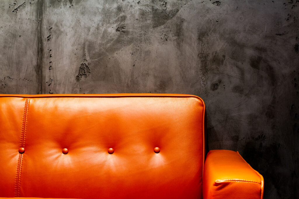 Ein orangefarbenes Ledersofa steht vor einer Betonoptik Wand, ideal für kreative Wandgestaltung im Wohnzimmer.